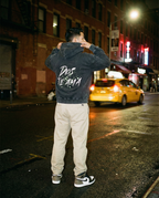 Person wearing a hoodie with text on the back, standing on a city street at night.